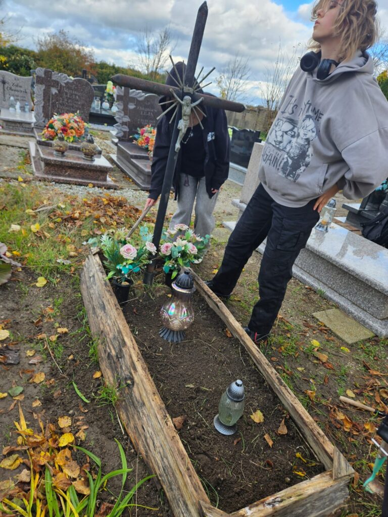 Ocalić od zapomnienia, czyli sprzątamy opuszczone groby. Na zdjęciu uporządkowany grób, na którym stoją dwa znicze. Pod krzyżem znajdują się sztuczne kwiaty. Z prawej strony stoi wolontariuszka, która oczyściła nagrobek