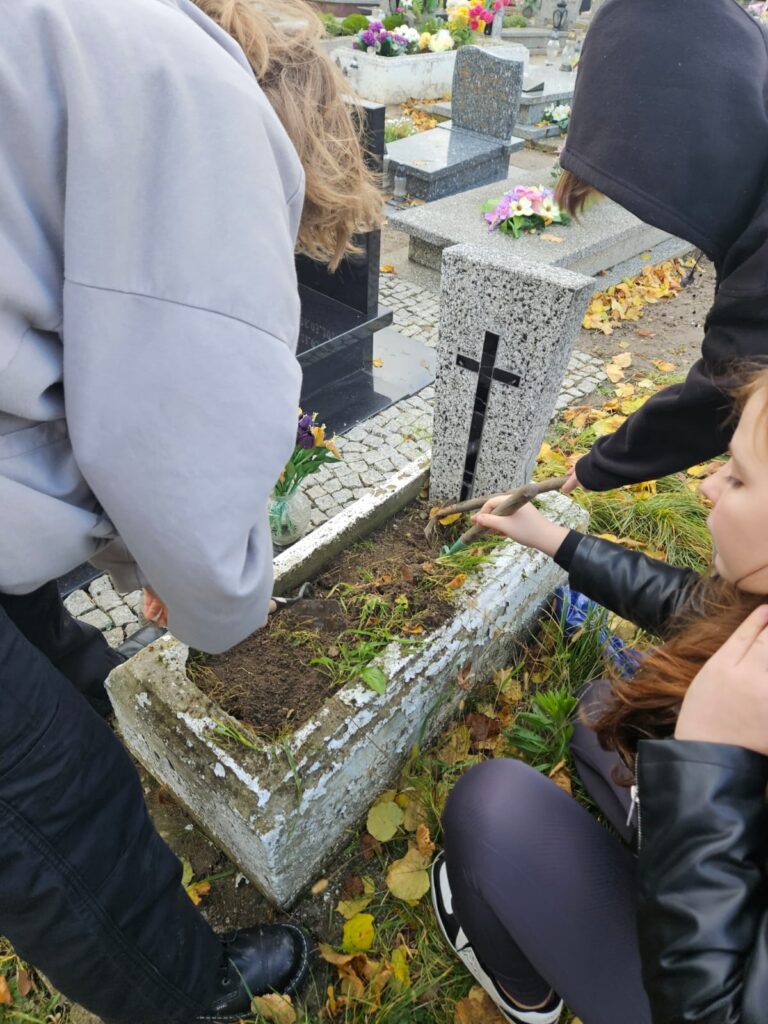 Ocalić od zapomnienia, czyli sprzątamy opuszczone groby. Na zdjęciu mały, stary nagrobek. Płyta nagrobna jest dość nowa. Z lewej strony wolontariuszka wyrywa trawę i chwasty. Z prawej strony klęcząca wolontariuszka, która usuwa chwasty z ziemi na nagrobku.