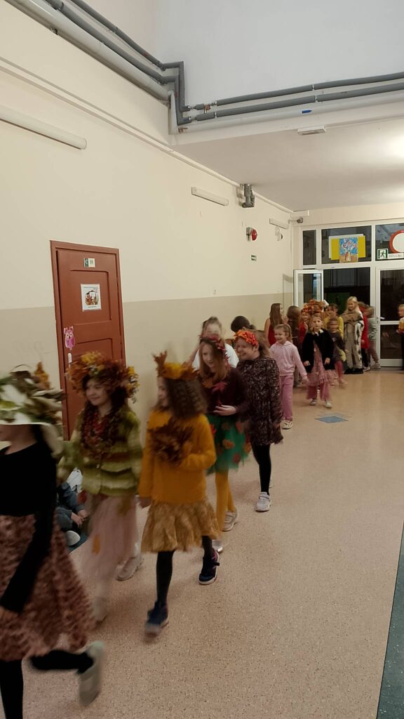 Dzień Dziewczynek w klasach 1-3. Na zdjęciu dziewczynki z spacerują po korytarzu szkolnym jak na wybiegu. Ich ubrania mają kolory jesieni: żółć, brąz, pomarańczowy. Na twarzach widać uśmiech.