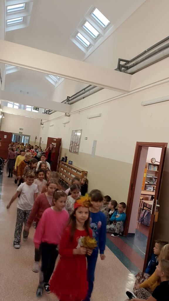 Dzień Dziewczynek w klasach 1-3. Na zdjęciu dziewczynki z spacerują po korytarzu szkolnym jak na wybiegu. Ich ubrania mają kolory jesieni: żółć, brąz, pomarańczowy. Na twarzach widać uśmiech.