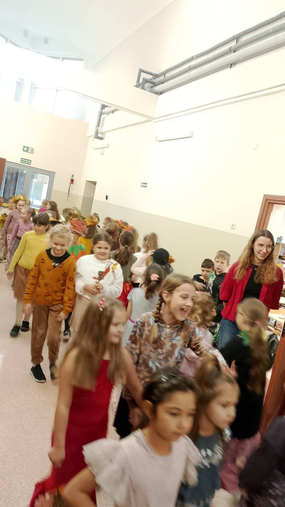 Dzień Dziewczynek w klasach 1-3. Na zdjęciu dziewczynki z spacerują po korytarzu szkolnym jak na wybiegu. Ich ubrania mają kolory jesieni: żółć, brąz, pomarańczowy. Na twarzach widać uśmiech. Po prawej stronie w drzwiach stoi nauczycielka z uśmiechniętą buzią.