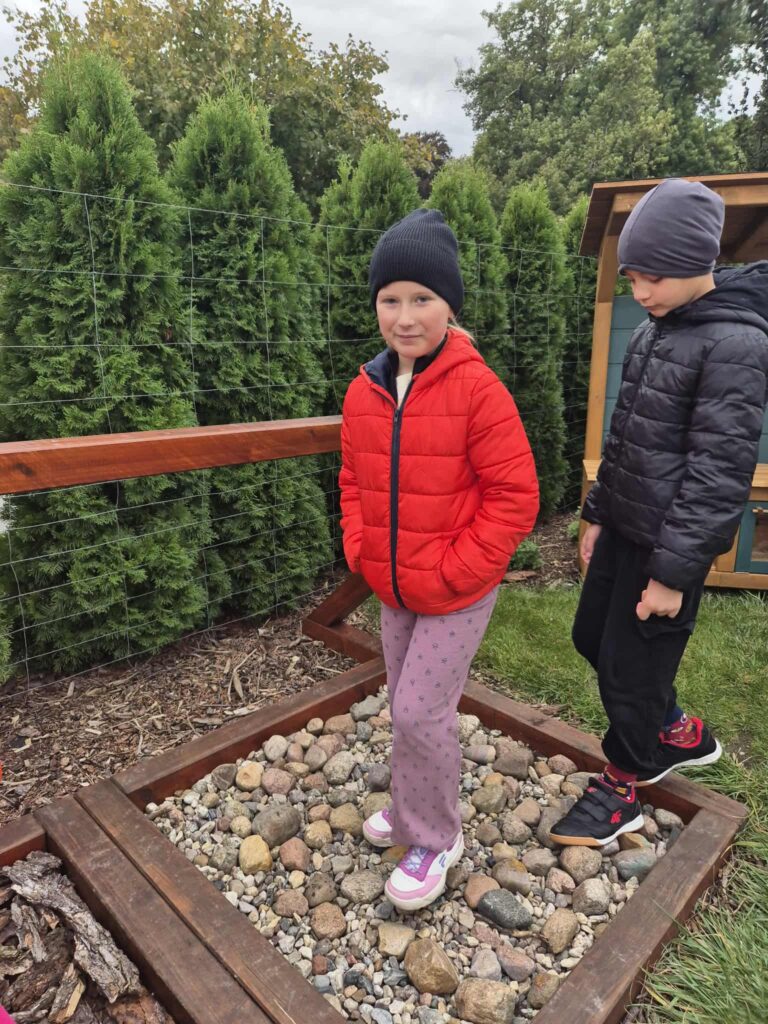 Wycieczka edukacyjna do Dalkowa. Na zdjęciu dwoje uczniów: dziewczynka i chłopiec spacerują po ścieżce sensorycznej ułożonej kamieniami i korą. W tle widać zielone drzewa