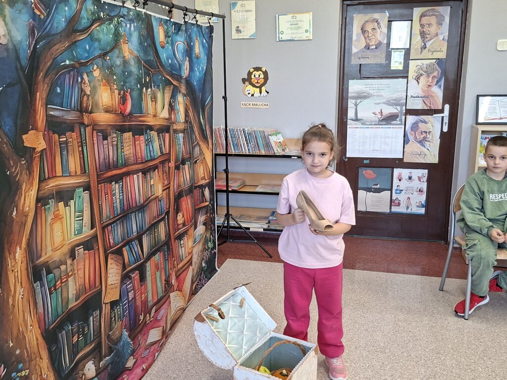 Przedstawia dziewczynkę trzymającą w ręce pantofelek. Obok niej kuferek z rekwizytami z baśni.
