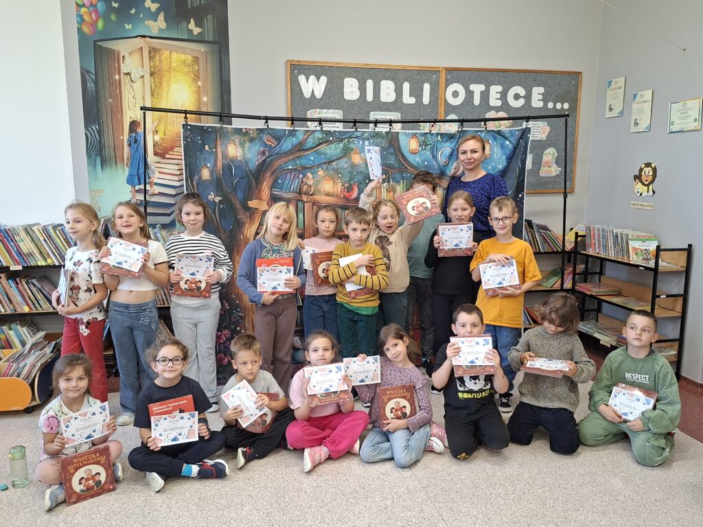 Przedstawia uczniów kl. 1c w bibliotece. 1 rząd uczniów siedzi, drugi stoi. Wszyscy trzymają dyplomy i książki. 
