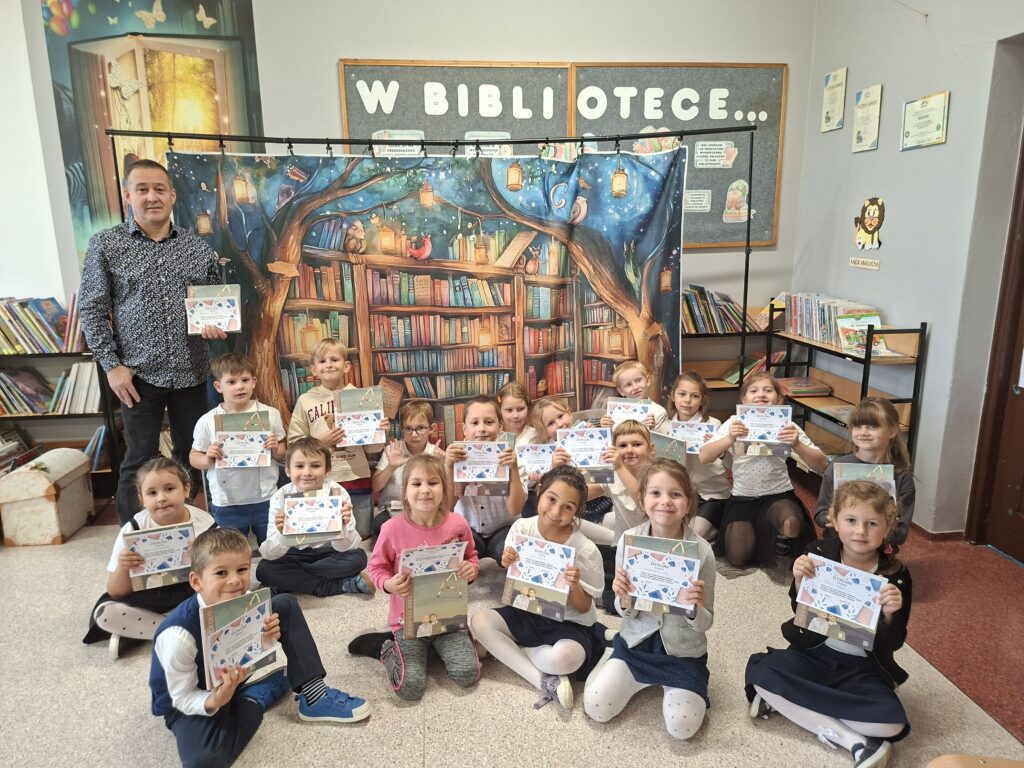 Przedstawia uczniów klasy 1a trzymających dyplom i pamiątkową książkę.  Za uczniami tło z książkami  i wiszącymi lampionami.