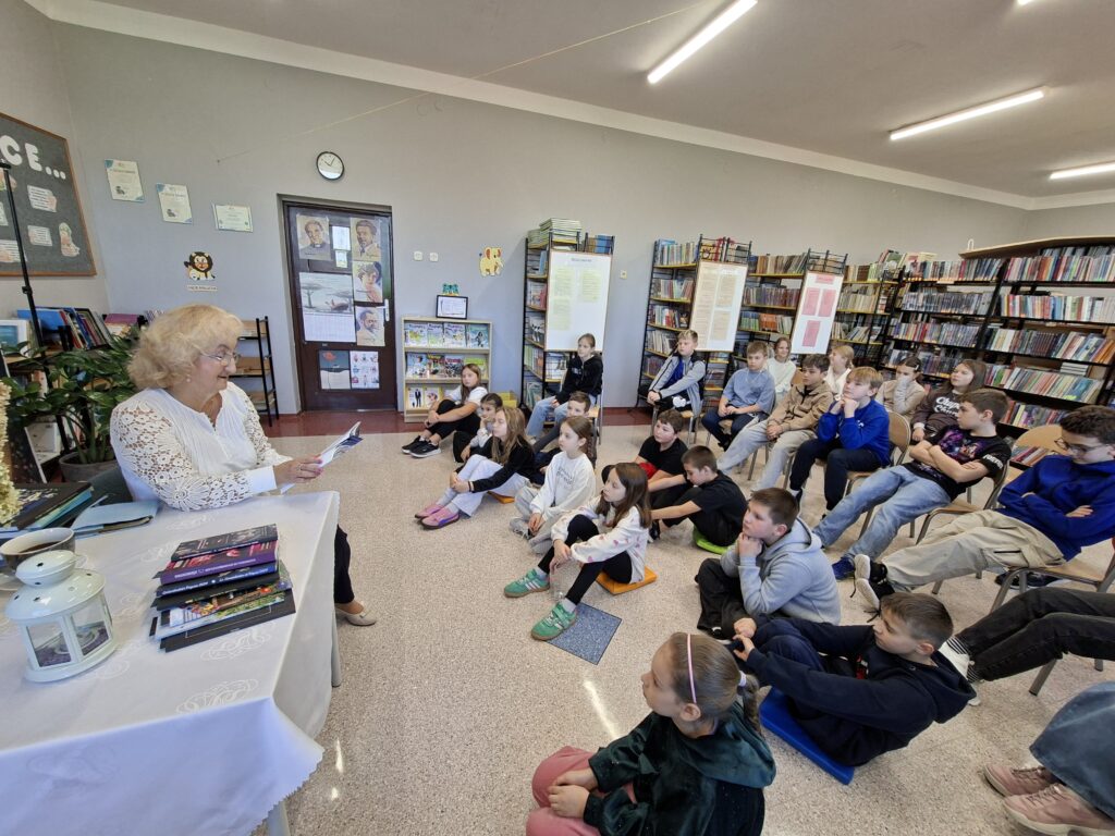 Przedstawia poetkę w białej bluzce siedzącą przy stoliku nakrytym białym obrusem. Na stole są tomiki wierszy poetkę i biała latarnia. Dzieci siedzą na poduszkach i krzesłach. 
