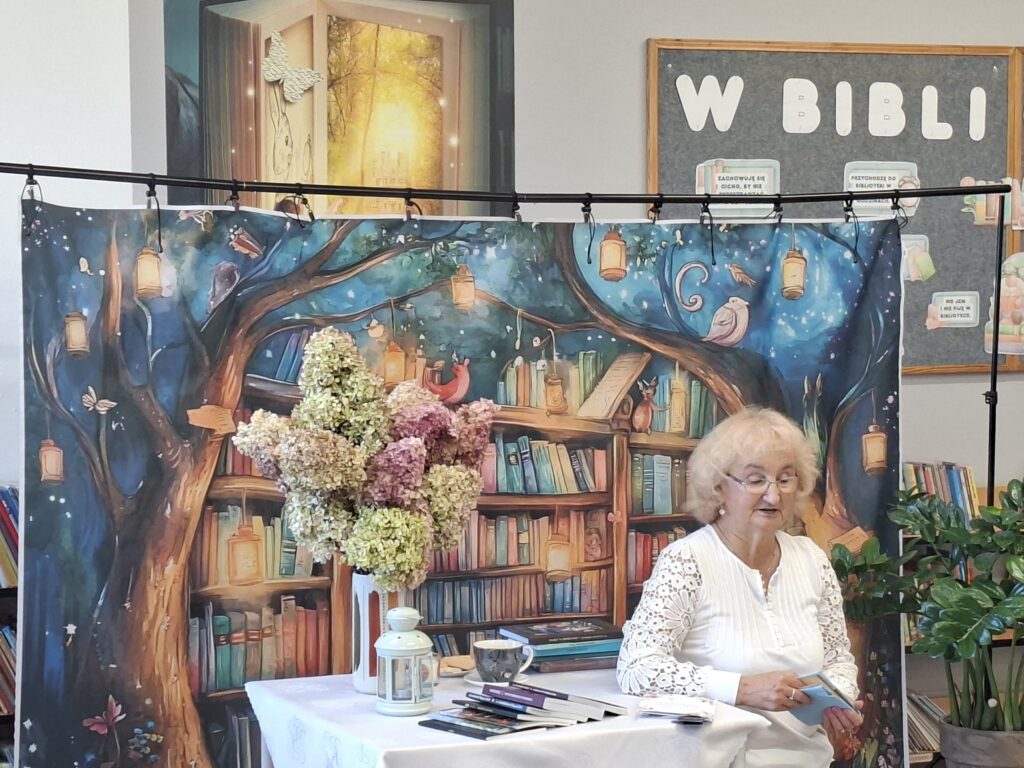 Zdjęcie przedstawia poetkę czytającą wiersze. Na stoliku hortensje w wazonie i biała latarnia. W tle książki na półkach.