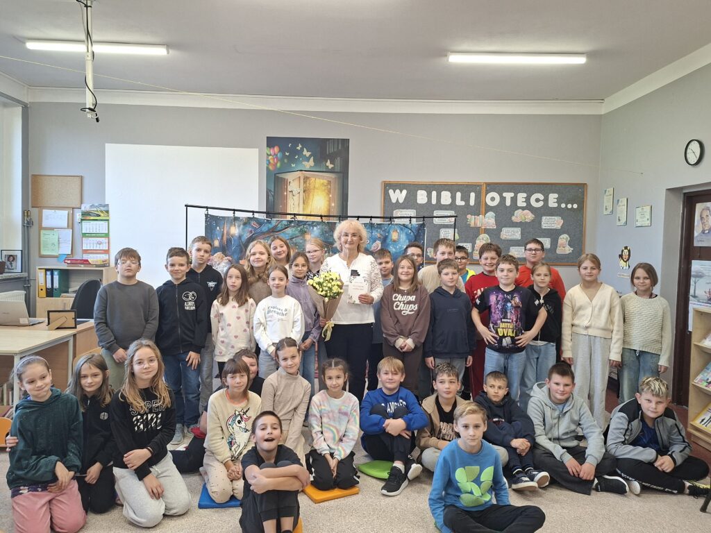 Zdjęcie przedstawia poetkę trzymająca żółte goździki i stojąca wokół uczniów klas 4. W tle napis w bibliotece..