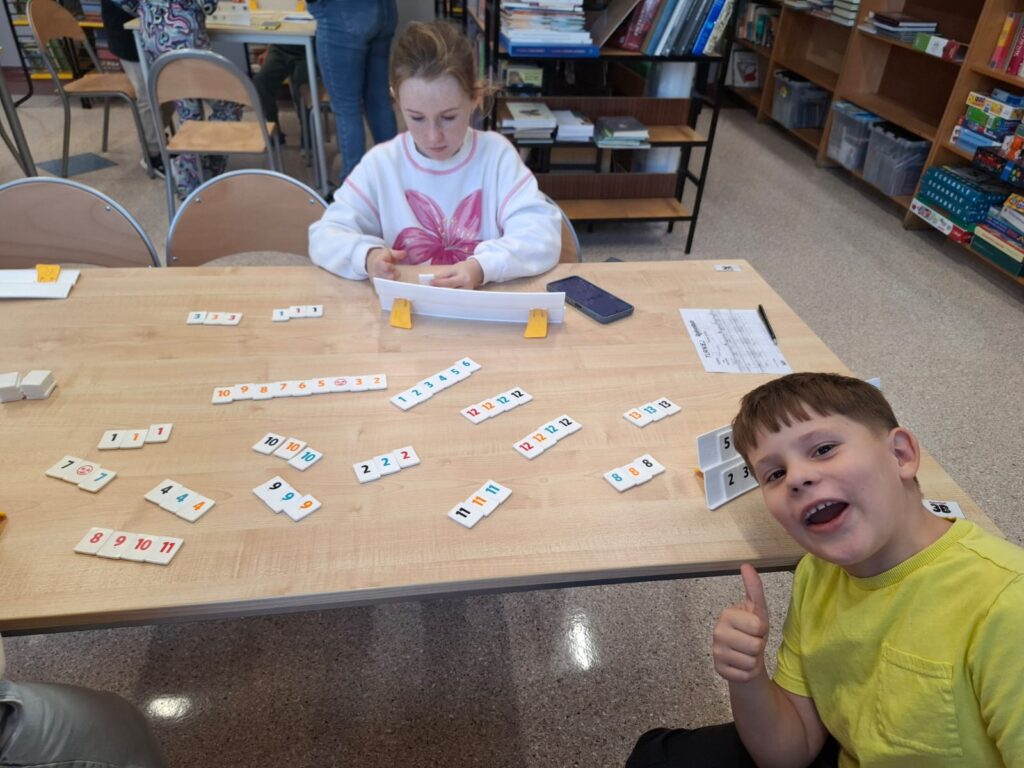 Zdjęcie przedstawia chłopca z uniesionym kciukiem w górę oraz dziewczynkę układająca kostki z liczbami na podstawce gry Rummikub.