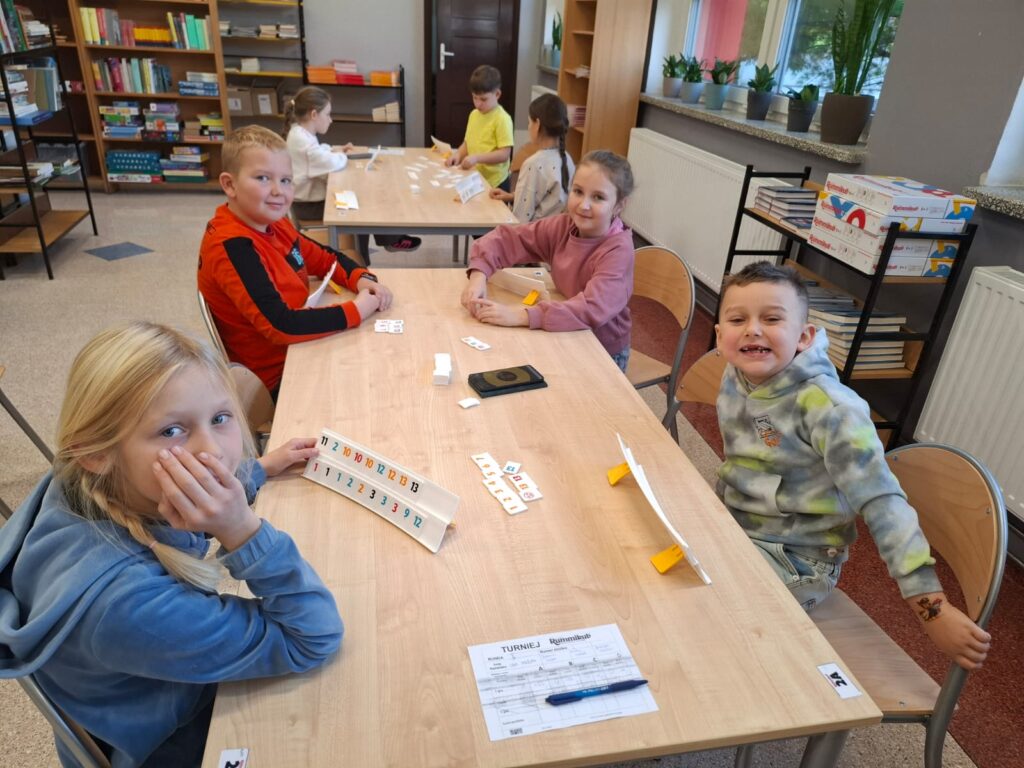 Zdjęcie przedstawia 4 uczniów siedzących przy stole i grających w Rummikub. Dzieci są uśmiechnięte.  Na stole są tabliczki z cyframi, długopis i kartka do zapisywania punktów. 