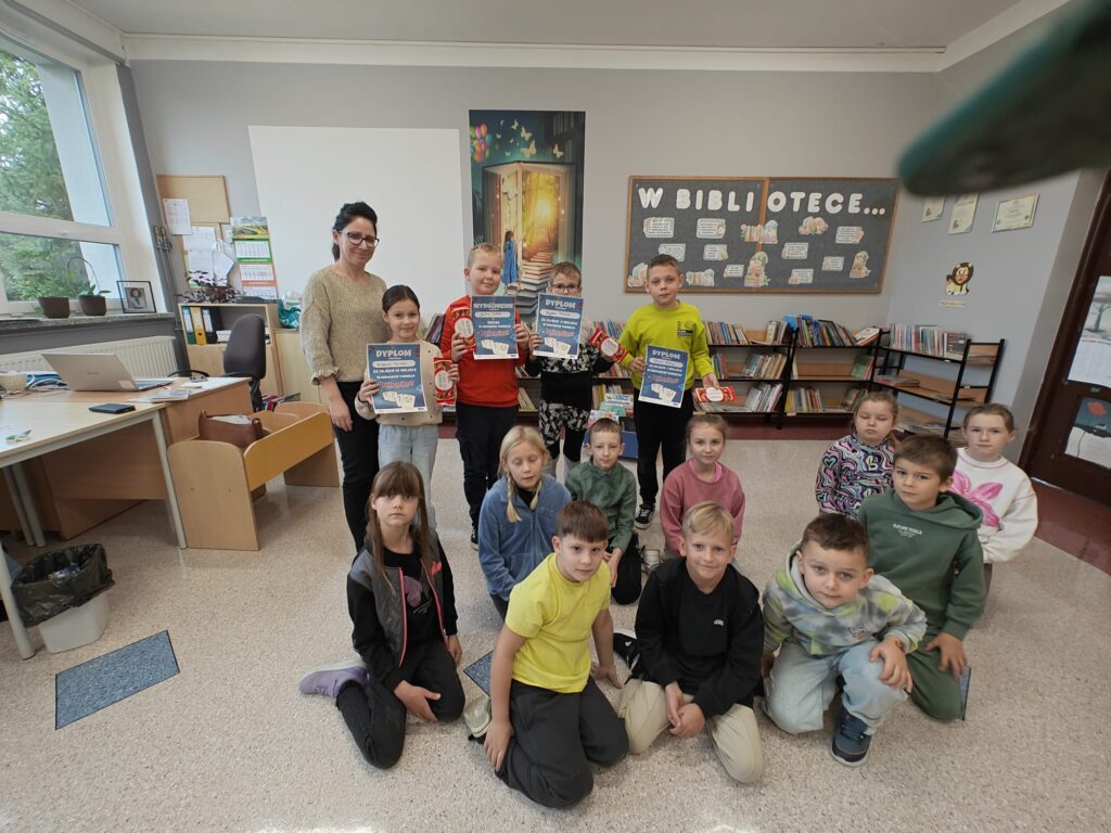 Zdjęcie przedstawia wszystkich uczestników turnieju, natomiast najlepsi z nich trzymają w rękach dyplomy I czekolady. W tle napis " W bibliotece...".