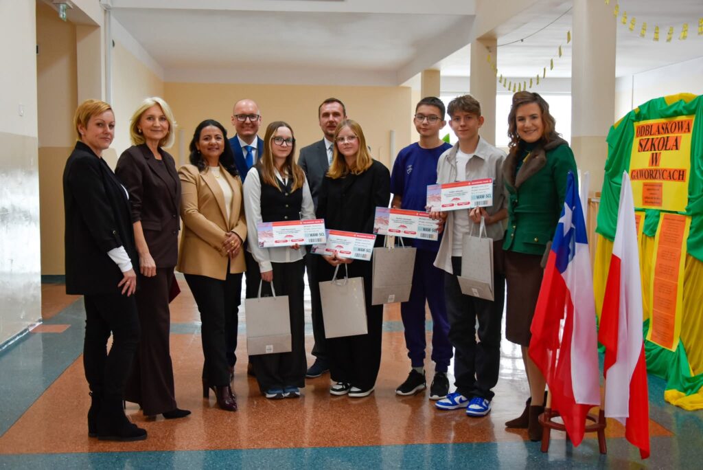 Uroczyste wręczenie biletów do Chile  na kolejną wymianę młodzieży. Na zdjęciu uczniowie, pani dyrektor, zastępca wójta i przedstawiciele Zarządu Sierra Gorda SCM z Chile pozują do zdjęcia. Uczniowie trzymają w rękach bilet. 
