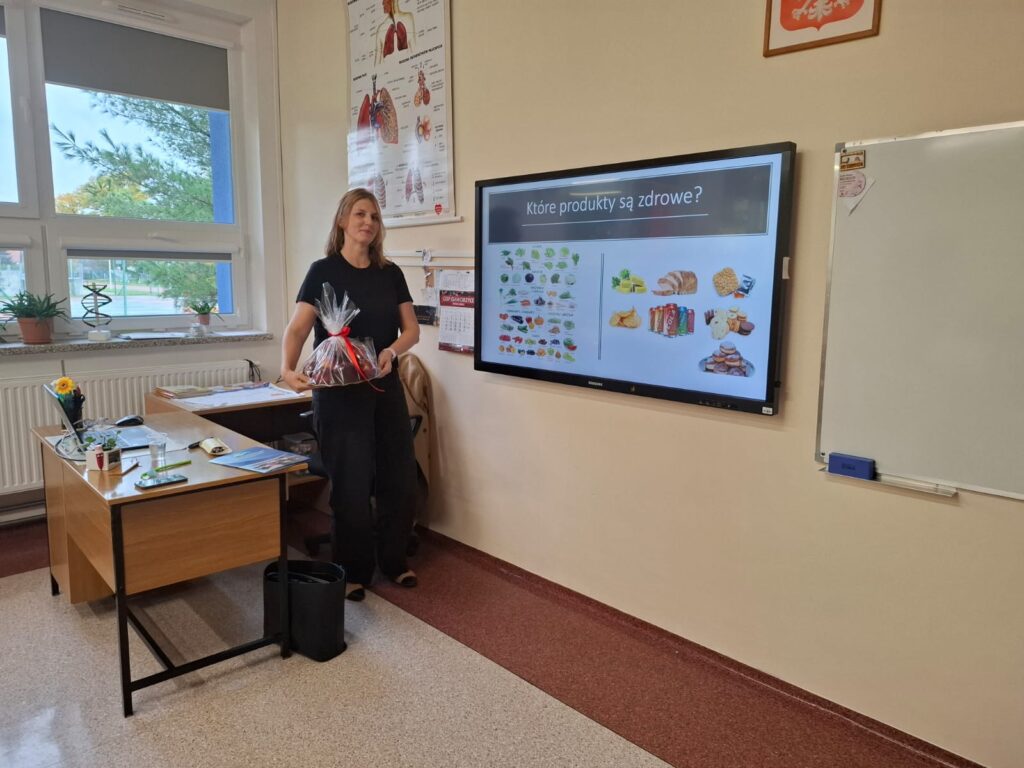 Spotkania ze specjalistami w ramach zajęć z edukacji zdrowotnej i przyrodniczej. Na zdjęciu jest pani dietetyk, trzyma upominek w postaci jabłek, po prawej stronie wyświetla się prezentacja na temat zdrowych produktów.