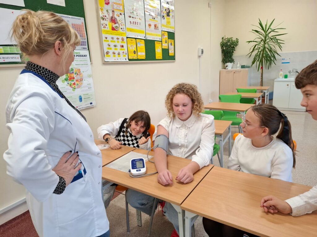 Spotkania ze specjalistami w ramach zajęć z edukacji zdrowotnej i przyrodniczej. Na zdjęciu trzy uczennice i szkolna pielęgniarka. Jedna z uczennic ma mierzone ciśnienie.