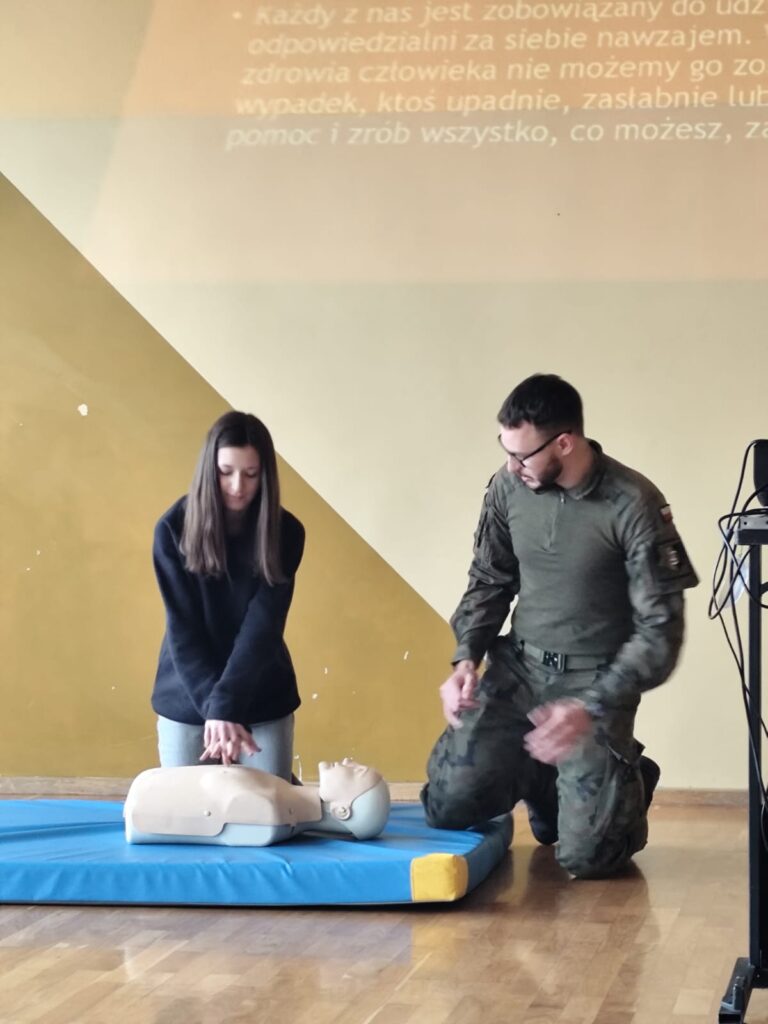 Edukacja z wojskiem. Na zdjęciu uczennica ćwiczy masaż serca na fantomie. Po jej lewej stronie jest żołnierz, który nadzoruje przebieg ćwiczenia.