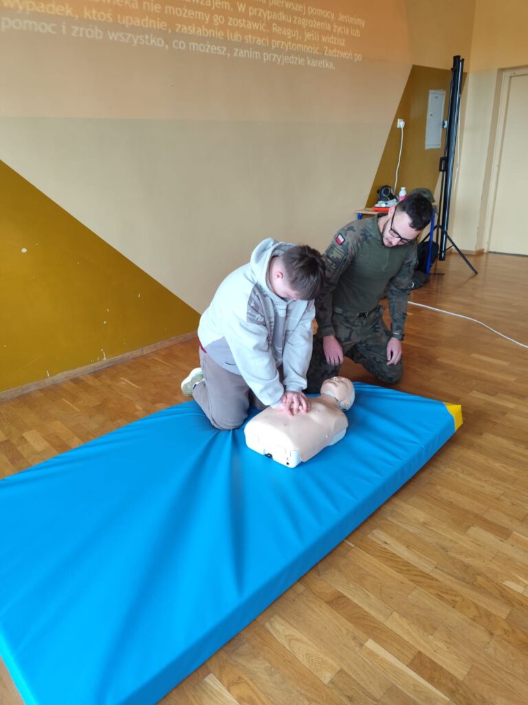 Edukacja z wojskiem. Na zdjęciu uczeń ćwiczy masaż serca na fantomie. Po jego lewej stronie jest żołnierz, który nadzoruje przebieg ćwiczenia.