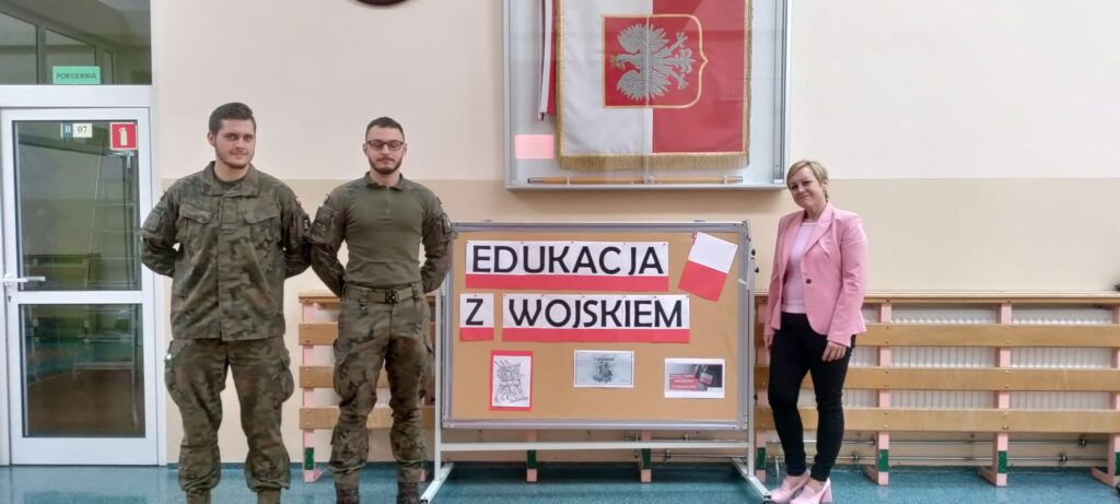 Edukacja z wojskiem. Na zdjęciu dwóch żołnierzy i pani Dyrektor stoją prze dekoracji z napisem "Edukacja z wojskiem".