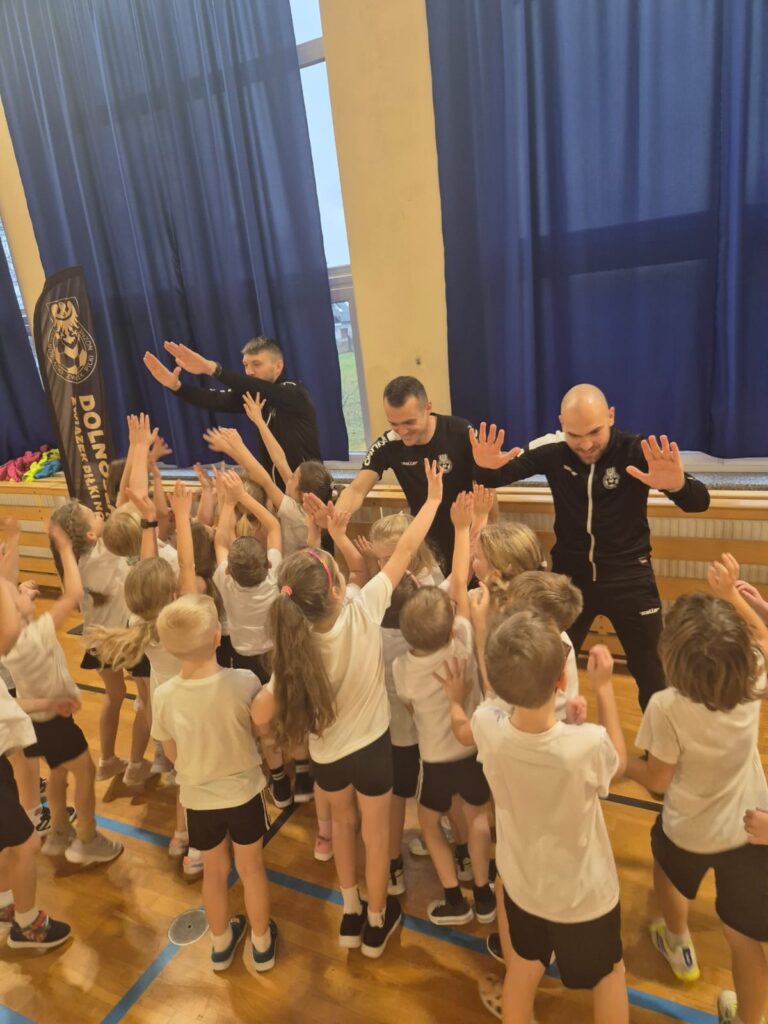 zDolnośląskie Dzieciaki. Na Zdjęciu uczniowie klasy pierwszej ubrani na sportowo w białe podkoszulki i czarne spodenki przybijają "piątkę"  trenerom piłki nożnej. 