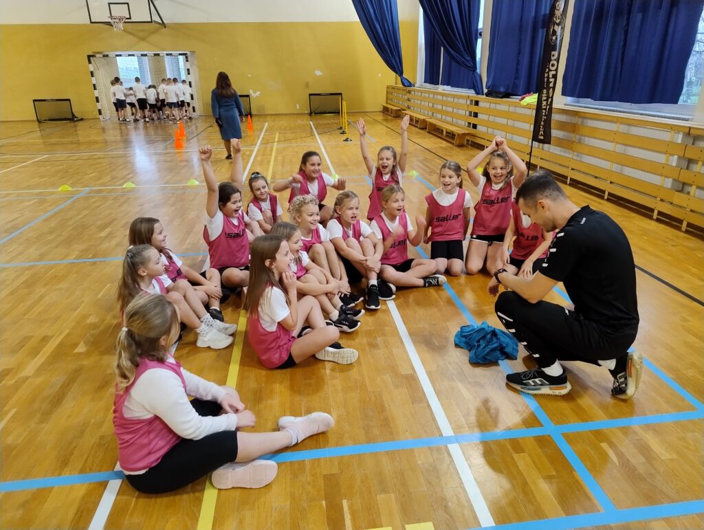 zDolnośląskie Dzieciaki. Dziewczynki siedzą na podłodze w sali gimnastycznej, mają na sobie kamizelki w kolorze ciemnego różu. Przed nimi kuca jeden z trenerów piłki nożnej. 