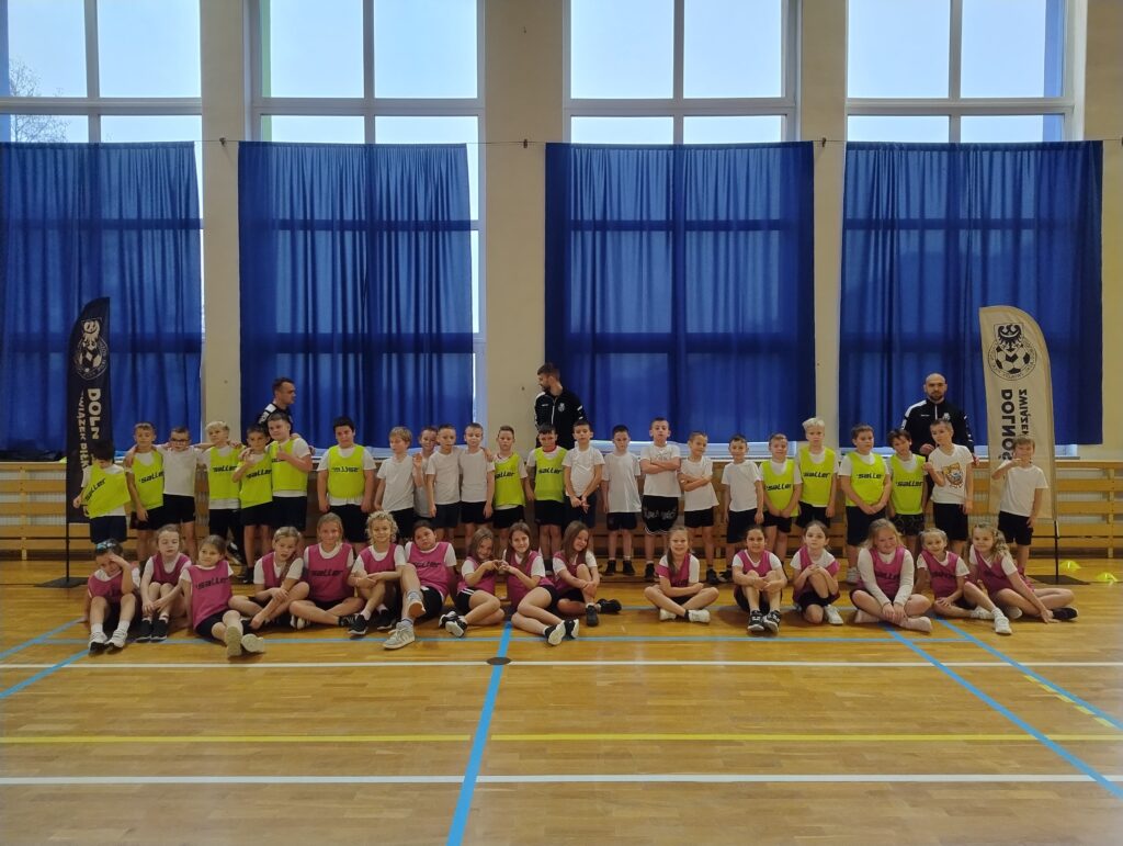 zDolnośląskie Dzieciaki. Na zdjęciu uczniowie klas drugich pozują do zdjęcia z trenerami. Chłopcy stoją pod oknem w jednym rzędzie, dziewczynki siedzą na podłodze.  