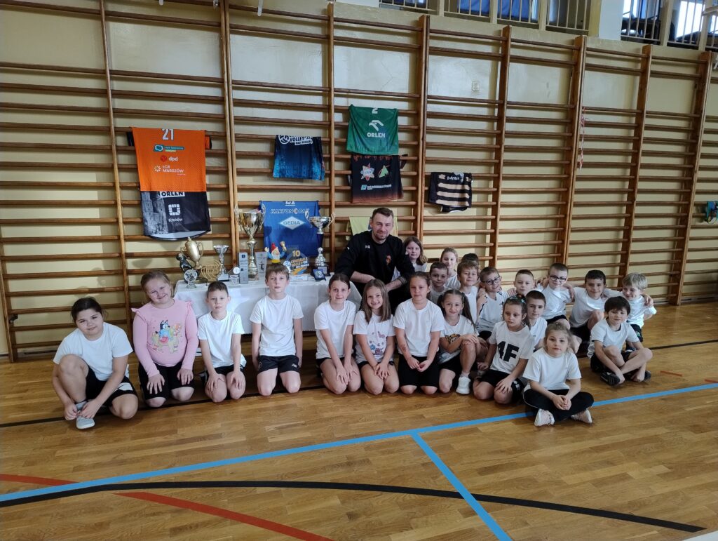 Zdjęcie dzieci na tle drabinek gimnastycznych, przedstawiające dzieci z trenerem siatkówki.
Za uczniami widać puchary, statuetki i medale oraz koszulki klubowe umieszczone na drabinkach.