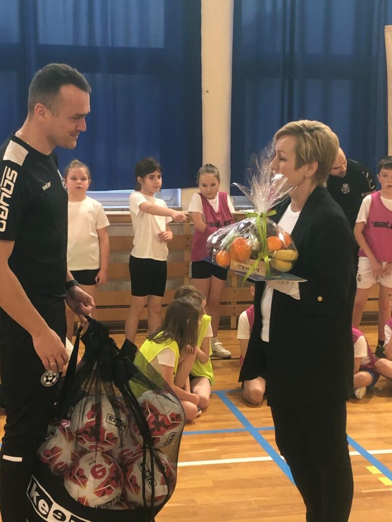 zDolnośląskie Dzieciaki. Na zdjęciu pani Dyrektor wręcza podziękowanie trenerom. W ręce trzyma zawinięte w celofanie owoce. Trener trzyma w ręce worek z piłkami do przekazania uczniom. 