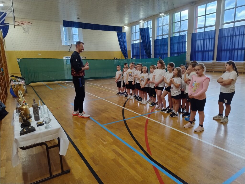 Zdjęcie przedstawia dzieci ustawione w szeregu przed trenerem siatkówki, który tłumaczy dzieciom zasady gry.