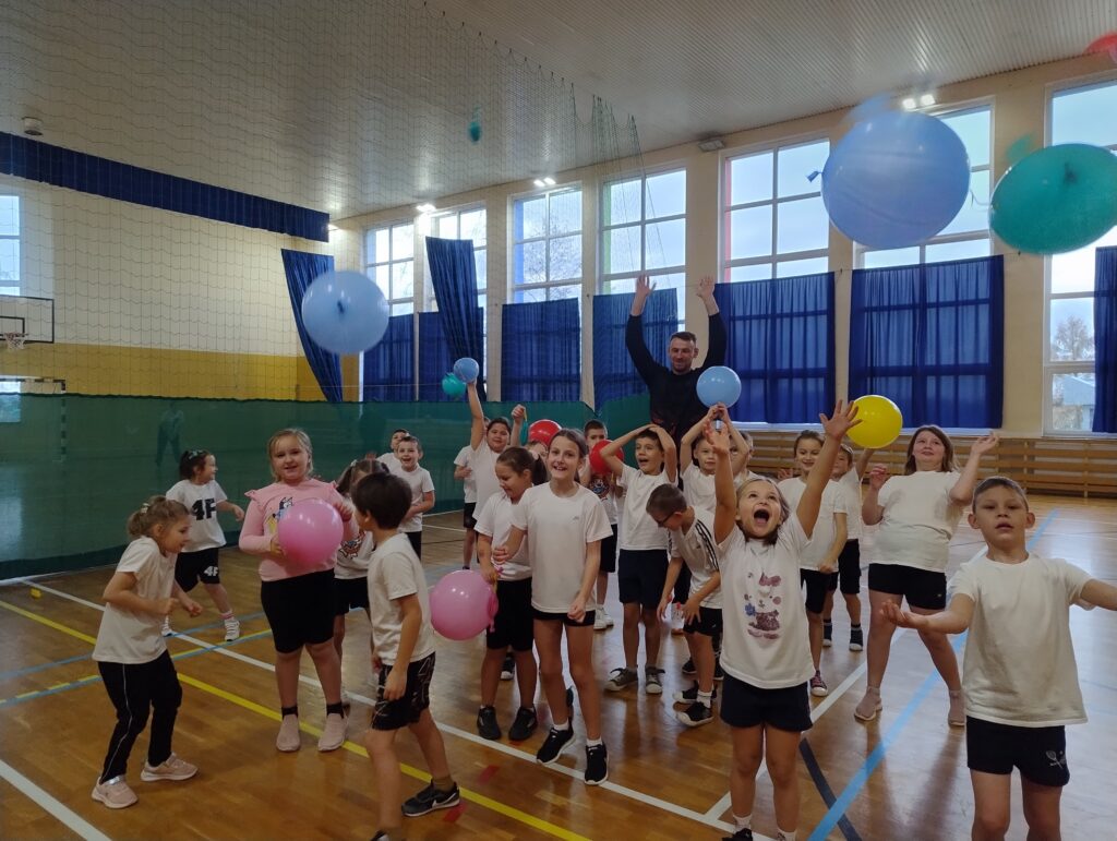 Zdjęcie przedstawia uśmiechnięte dzieci, które podrzucają kolorowe balony do góry. Dzieci ćwiczą z trenerem na sali gimnastycznej.