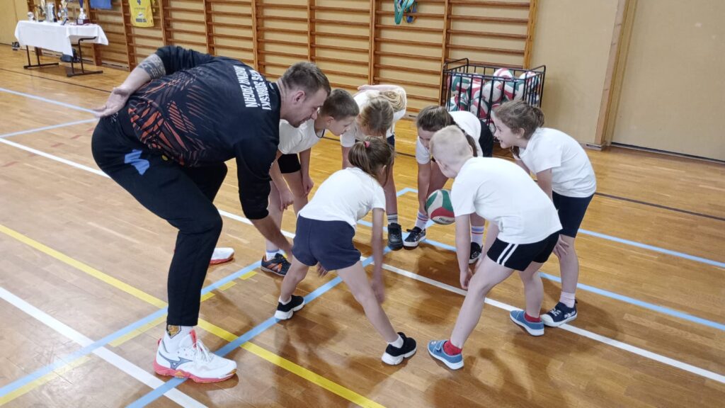 Dzieci stoją w kole ustawione do zabawy z trenerem, są pochylone i zwrócone twarzą do środka koła.
Przetaczają do siebie piłkę do siatkówki po podłodze.