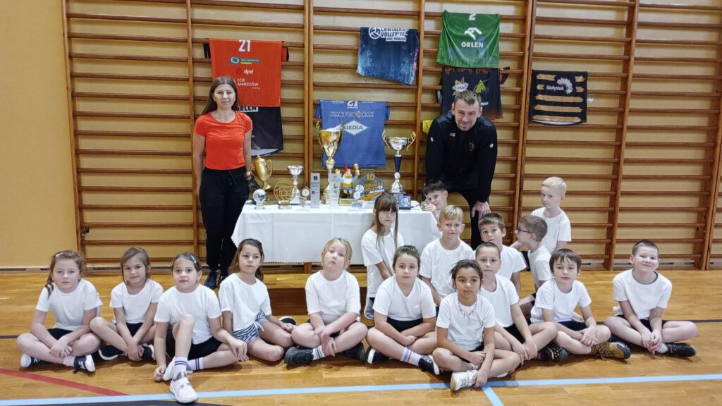 Zdjęcie grupowe na tle drabinek gimnastycznych, przedstawiające dzieci z trenerem siatkówki oraz nauczycielką w-f.
Za uczniami widać wyeksponowane na stoliku puchary, statuetki i medale oraz koszulki klubowe umieszczone na drabinkach.