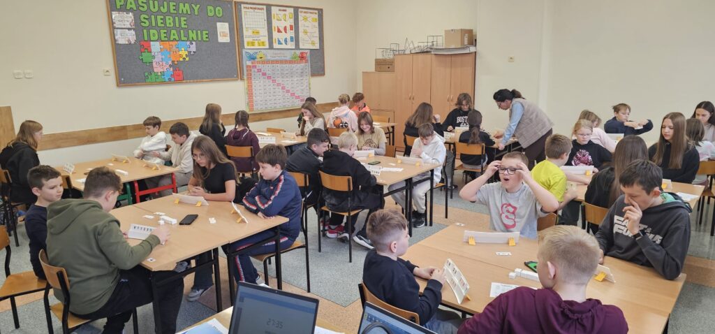 Na zdjęciu widzimy 35 uczniów przy stolikach 4-osobowych, uczniowie grają w Rummikuba, na stolikach rozłożone są kostki Rummikuba. Przy jednym stanowisku jest nauczyciel, który nadzoruje przebieg gry.