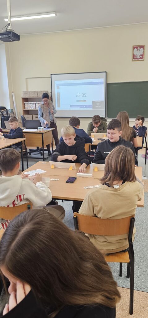 Zdjęcie przedstawia widok dwóch stolików z uczniami grającymi w Rumikuba, w tle stoi nauczyciel nadzorujący grę, widać tablicę multimedialną z mierzonym czasem jednej rundy.