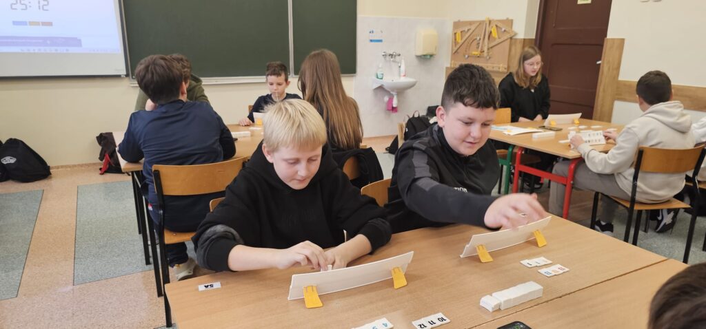 Zdjęcie przedstawia widok panoramiczny całej klasy z dziewięcioma stolikami, przy każdym siedzi czterech uczniów grających w Rummikuba.