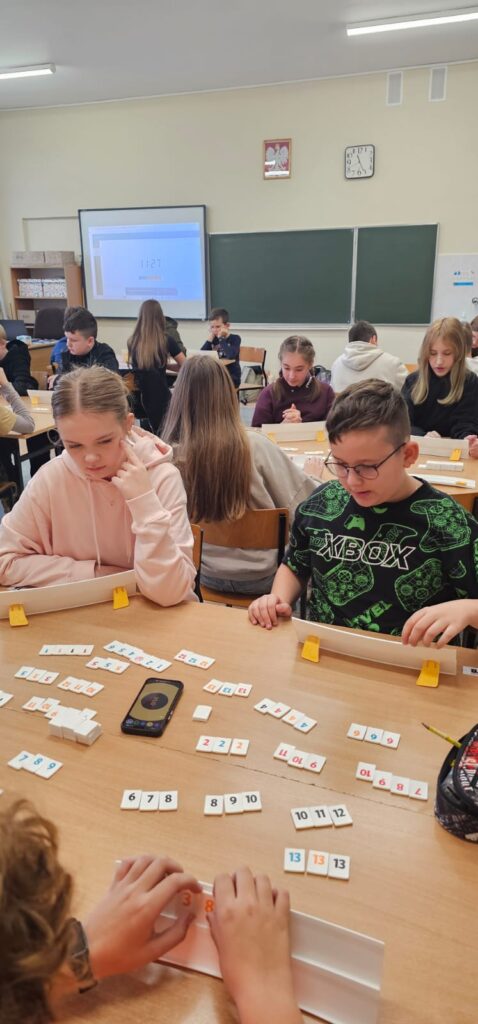 Zdjęcie przedstawia zbliżenie dwóch uczniów: dziewczynka i chłopiec, którzy grają w Rummikuba, w tle widać trzy pozostałe stoliki z zawodnikami i tablicę interaktywną.