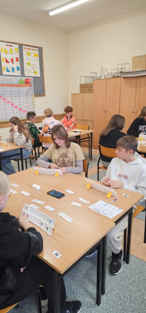 Zdjęcie przedstawia zbliżenie uczennicy i ucznia przy stoliku z klockami Rummikuba w trakcie gry, za nimi widać trzy pozostałe stoliki z zawodnikami.