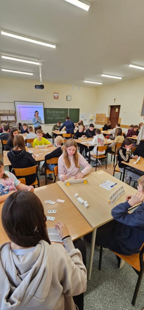 Zdjęcie przedstawia widok całej klasy z uczniami przy stoliku, którzy grają w Rummikuba, przy biurku stoi nauczyciel, który informuje uczestników, ile czasu pozostało do końca rundy, za nauczycielem widać tablicę interaktywną z zegarem odmierzającym czas.