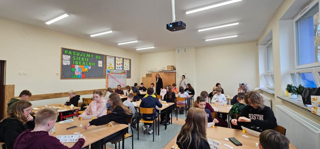  Zdjęcie przedstawia widok panoramiczny całej klasy ze wszystkimi uczniami przy stolikach, którzy grają w Rummikuba oraz dwóch nauczycieli nadzorujących przebieg gry.