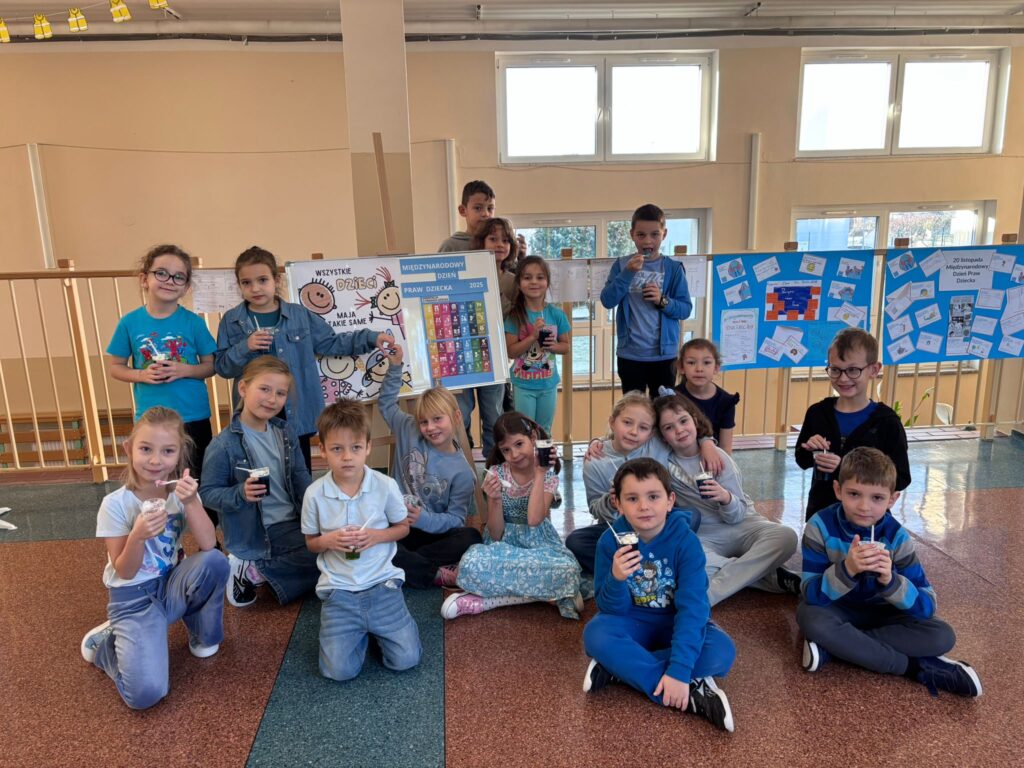 Światowy Dzień Praw Dziecka. Na zdjęciu znajduje się grupa dzieci z klasy 1c ubranych w niebieskie koszulki, siedzących i stojących razem w szkolnej przestrzeni. Uczniowie trzymają w rękach plastikowe kubki z niebieskimi galaretkami. W tle widać tablicę z niebieskimi plakatami oraz opisami, a także prace uczniów klas 4-8 i kartki przypięte do barierki.