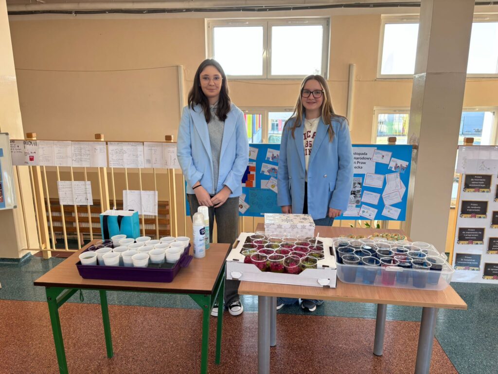 Światowy Dzień Praw Dziecka.  Na zdjęciu widać dwie uczennice, z dwoma stolikami ustawionymi w szkolnym korytarzu. Na stolikach znajdują się liczne plastikowe kubeczki z galaretkami. W tle widać tablicę z niebieskimi plakatami oraz opisami, a także prace i kartki przypięte do barierki.