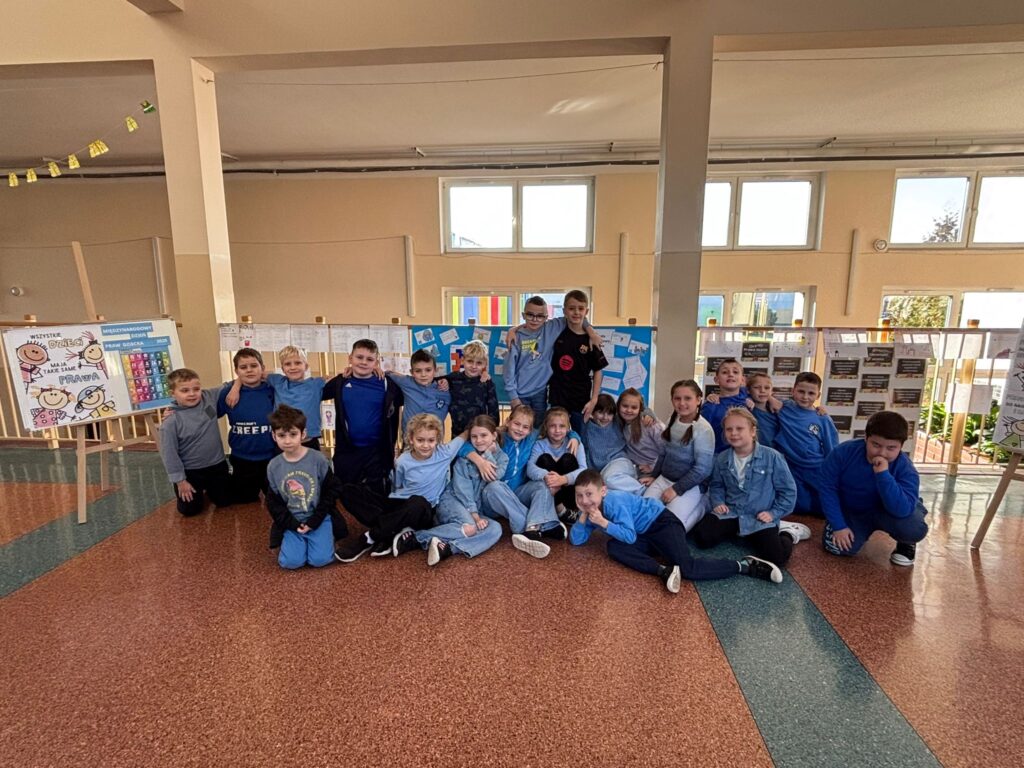 Światowy Dzień Praw Dziecka. Na zdjęciu znajduje się grupa dzieci z klasy 2b ubranych w niebieskie koszulki, leżących  i stojących razem w szkolnej przestrzeni. W tle widać tablicę z niebieskimi plakatami oraz opisami, a także prace uczniów klas 4-8 i kartki przypięte do barierki.