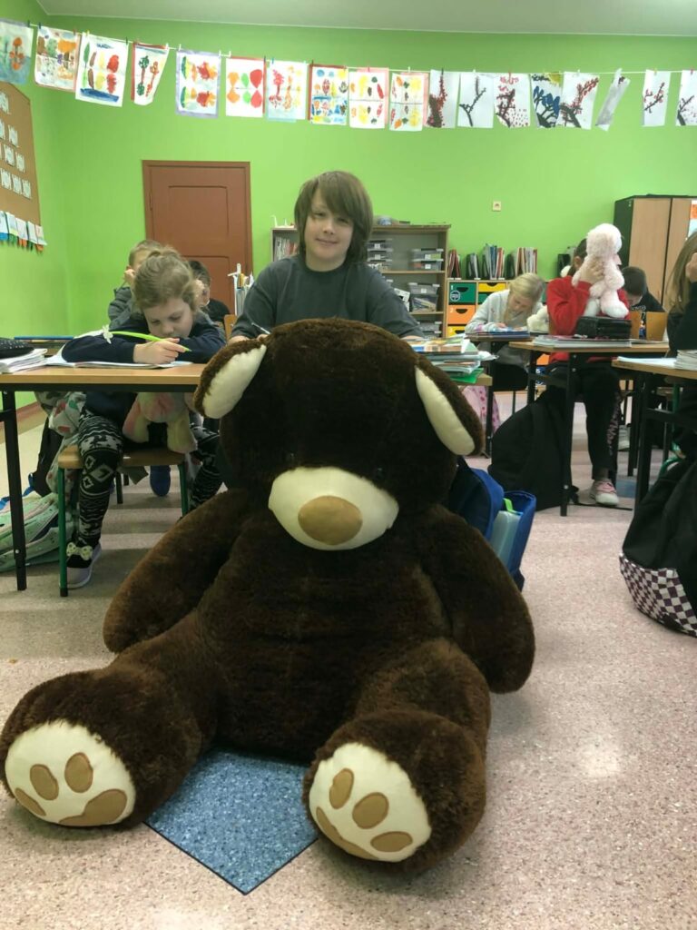 Na zdjęciu wielki brązowy miś siedzi na podłodze, za nim widać uczniów z innymi misiami. Brązowy miś Mikołaj – to miś jego babci.