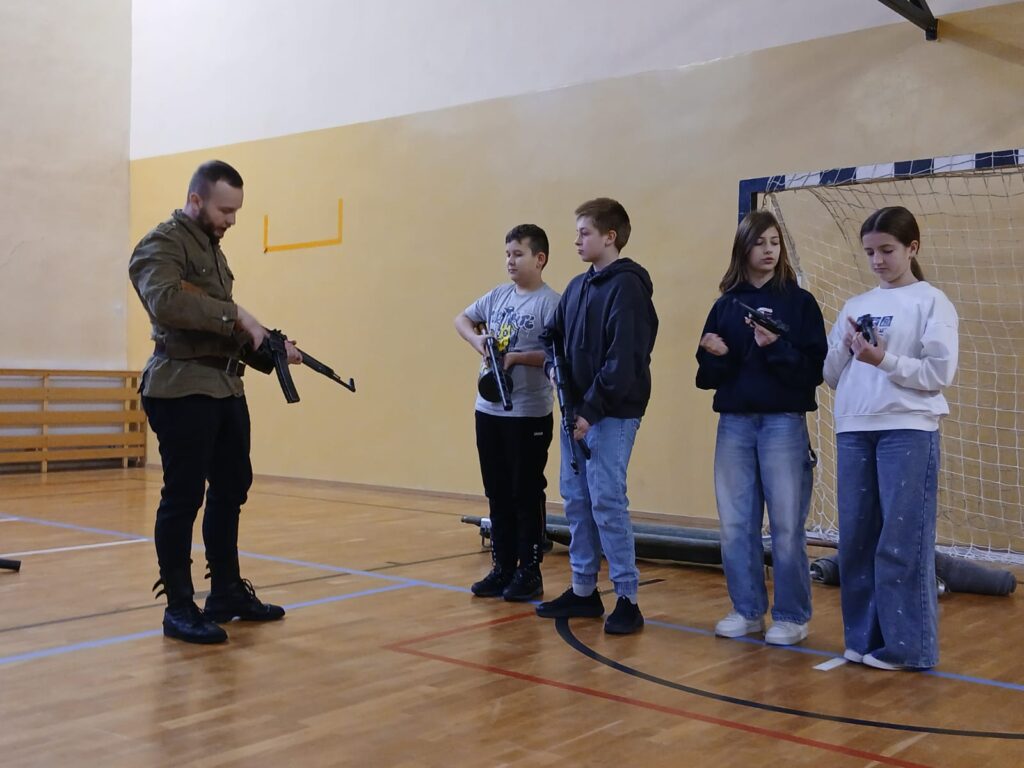 Na zdjęciu widać rekonstruktora z grupy „Rekonstrukto” prezentuje uczniom umundurowanie i wyposażenie żołnierzy Armii Krajowej. Trzyma replikę broni, a dzieci uważnie słuchając opowieści o Powstaniu Warszawskim.