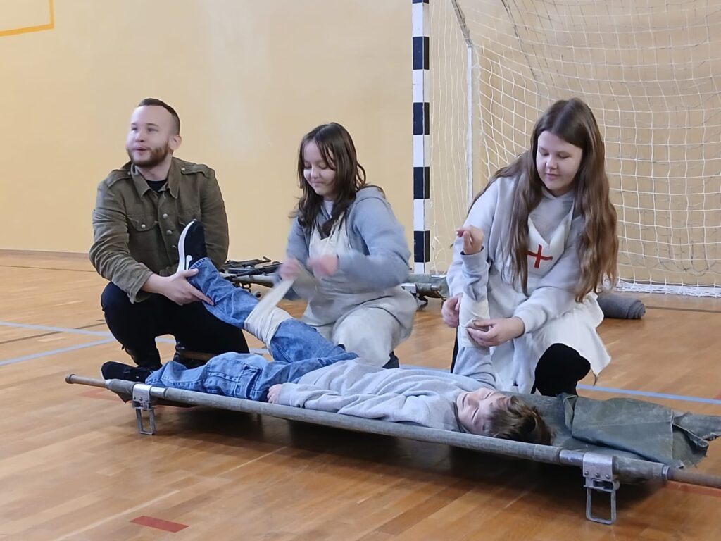 Na zdjęciu znajdują się dwie sanitariuszki w powstańczych strojach oraz rekonstruktor w mundurze żołnierza Armii Krajowej. Sanitariuszki – uczennice z klasy trzymają bandaże i udzielają pomocy rannemu powstańcowi leżącemu na noszach.