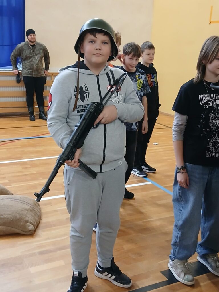 Na zdjęciu uczeń klasy 6 trzyma replikę broni używaną w czasie Powstania Warszawskiego. W tle widać zaciekawionych uczniów i jednego rekonstruktora.