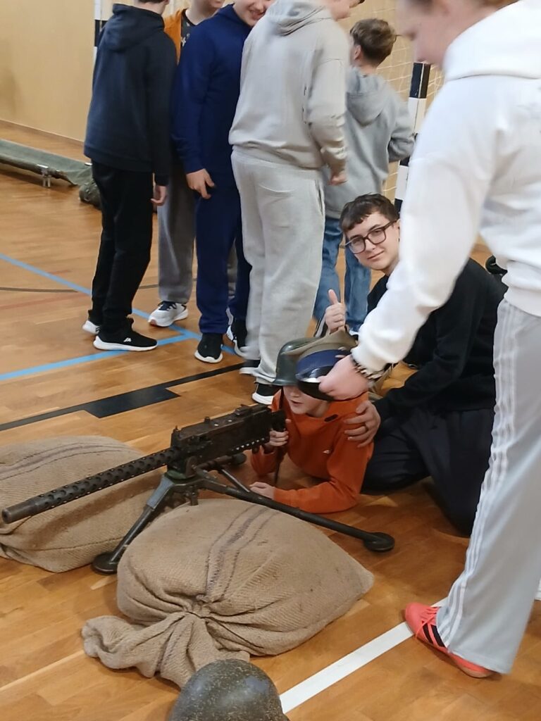 Na pierwszym planie zdjęcia widać stanowisko strzelnicze z repliką broni i ucznia, który mierzy z broni w pozycji leżącej . W tle widać grupę uczniów.