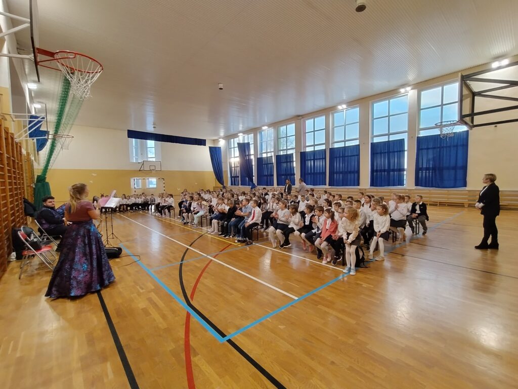 . Zdjęcie przedstawia kobietę w przepięknej, fioletowej sukni. Kobieta trzyma w dłoni mikrofon, przez który opowiada widowni historię o królewnie zaklętej w żabę. Na drugim tle widać dwóch siedzących mężczyzn. Mężczyzna po lewej stronie trzyma saksofon sopranowy, a drugi puzon.