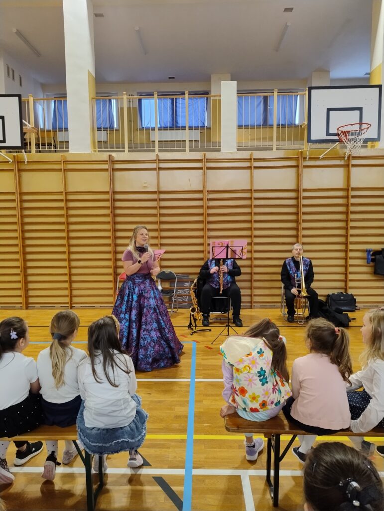 Zdjęcie przedstawia muzyków występujących przez publicznością, tj. uczniami klas 1-3. Uczniowie siedzą na drewnianych ławkach. Są ubrani w galowe stroje – białe koszule oraz ciemne spódniczki i spodnie.