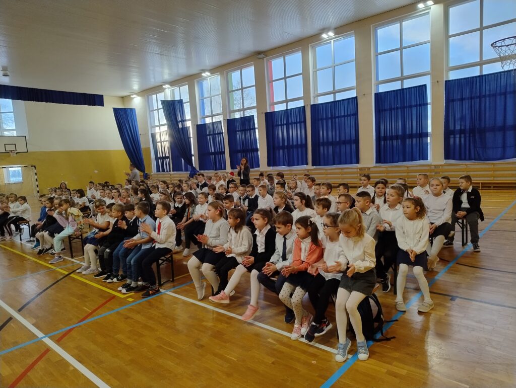 Zdjęcie pokazuje w przybliżeniu uczniów klas 1-3 siedzących na widowni w galowych strojach.