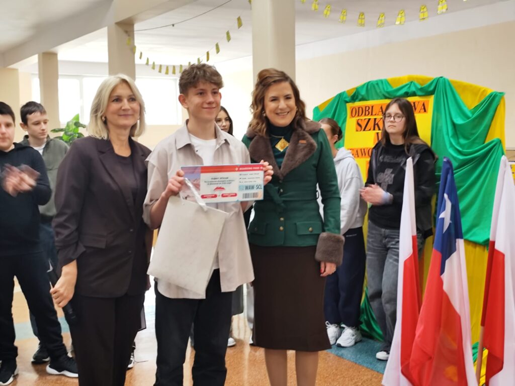 Uroczyste wręczenie biletów do Chile  na kolejną wymianę młodzieży. Na zdjęciu uczeń, który otrzymał bilet do Chile na wymianę młodzieży. Po prawej stronie dwie flagi Polski i jedna Chile. 