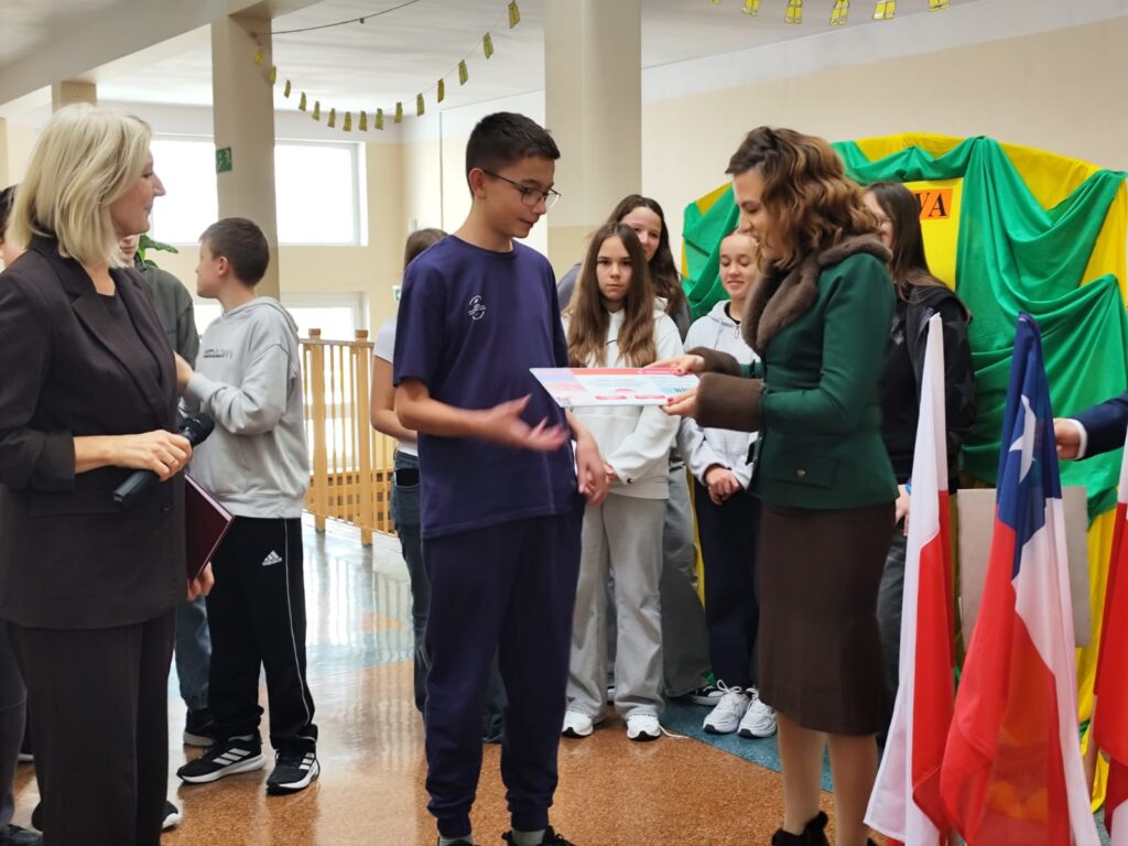 Uroczyste wręczenie biletów do Chile  na kolejną wymianę młodzieży. Na zdjęciu wiceprezes ds. prawnych Sierra Gorda SCM wręcza uczniowi bilet do Chile. W tle inni uczniowie. 
